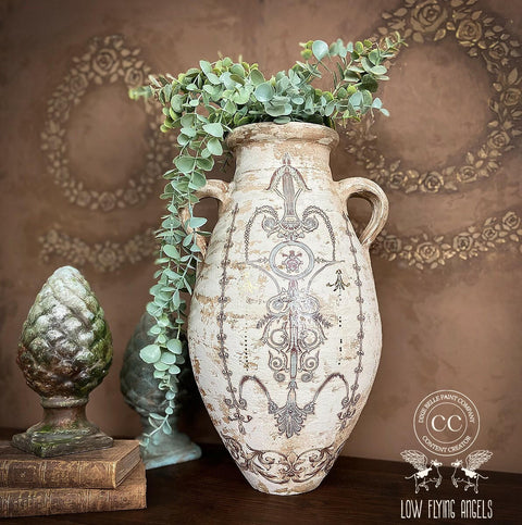 Large white distressed concrete vase with 2 handles and faux eucalyptus overflowing out of it. The Gilded Damask Transfer has been applied to the front. Staged next to a stack of books with a  patina'd sculpture of a pinecone on top.