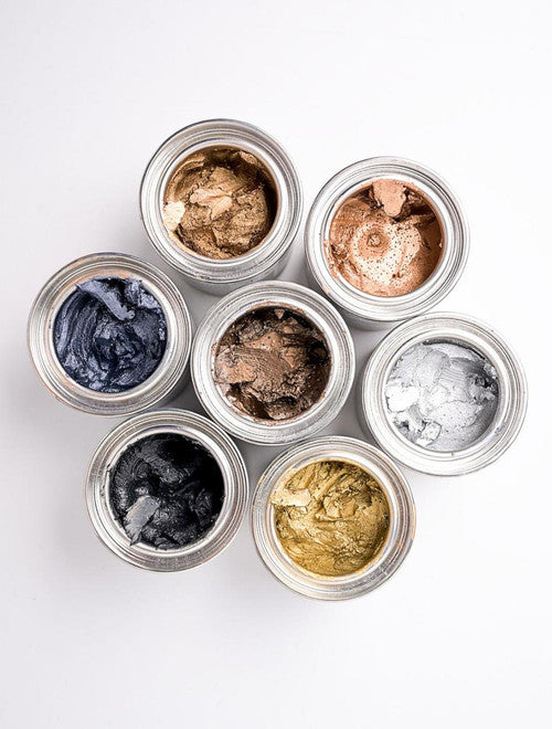 Seven open jars of metallic pigment waxes in gold, silver, bronze, black, and blue shades arranged in a flower-like pattern on a white background.