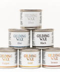 Six cans of gilding wax are stacked in a pyramid. Top row: Silver. Middle row: Zinc, Black. Bottom row: Copper, Gold, Bronze. Each can has a white label with detailed text around the name.