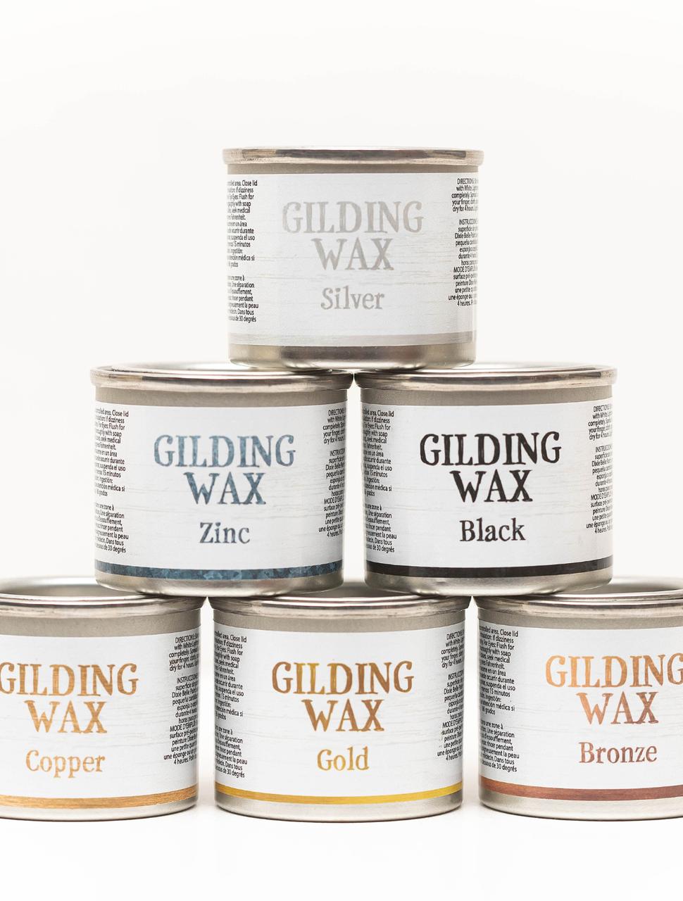 Six cans of gilding wax are stacked in a pyramid. Top row: Silver. Middle row: Zinc, Black. Bottom row: Copper, Gold, Bronze. Each can has a white label with detailed text around the name.