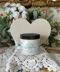 Grayish white painted wooden heart with a jar of Glacier in front of it. Staged on crocheted lace with faux greenery and flowers.