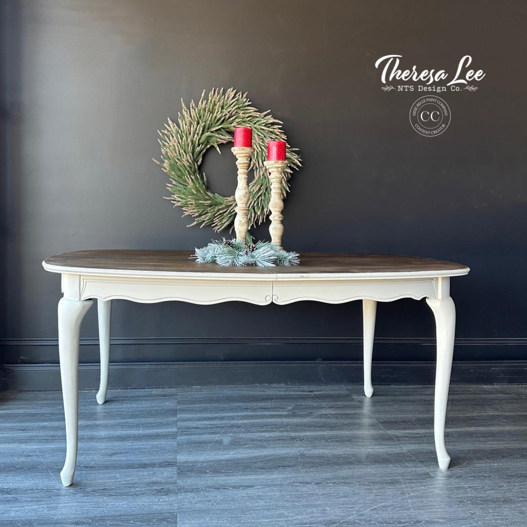 Grayish white painted coffee table with a medium wood top. Staged with candlesticks and garland on top. A green wreath hangs behind the piece on the wall.