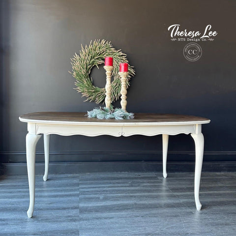 Grayish white painted coffee table with a medium wood top. Staged with candlesticks and garland on top. A green wreath hangs behind the piece on the wall.