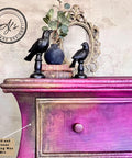 A vintage dresser with a gradient of purple and gold hues is adorned with two black bird figurines, a small vase with leaves, and stacked books. An ornate mirror is placed behind them. The image showcases the mix of Gold and Bronze Gilding wax.