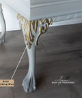 Close-up of a table leg painted in white with gold gilding wax on ornate details. The leg has a curved, decorative foot. A label points to the gold area, and the floor appears to be wooden. The image has the text "Bow Tie Treasures.
