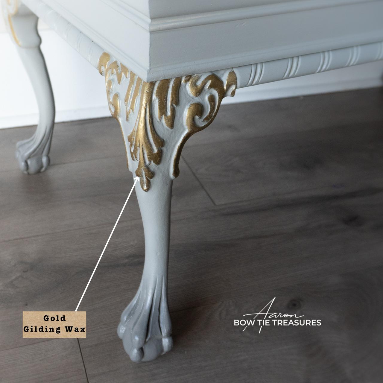 Close-up of a table leg painted in white with gold gilding wax on ornate details. The leg has a curved, decorative foot. A label points to the gold area, and the floor appears to be wooden. The image has the text "Bow Tie Treasures.
