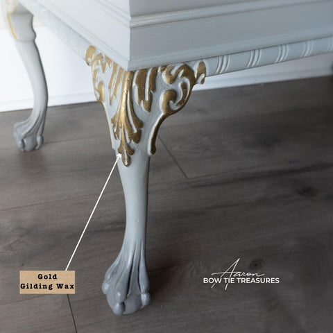 Close-up of a table leg painted in white with gold gilding wax on ornate details. The leg has a curved, decorative foot. A label points to the gold area, and the floor appears to be wooden. The image has the text "Bow Tie Treasures.