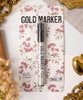 Packaging of a gold marker with a chisel tip. The card backing features a floral design in pink and white tones. The brand name "Belles and Whistles" is visible. Gold decorative elements and white fabric are in the background.