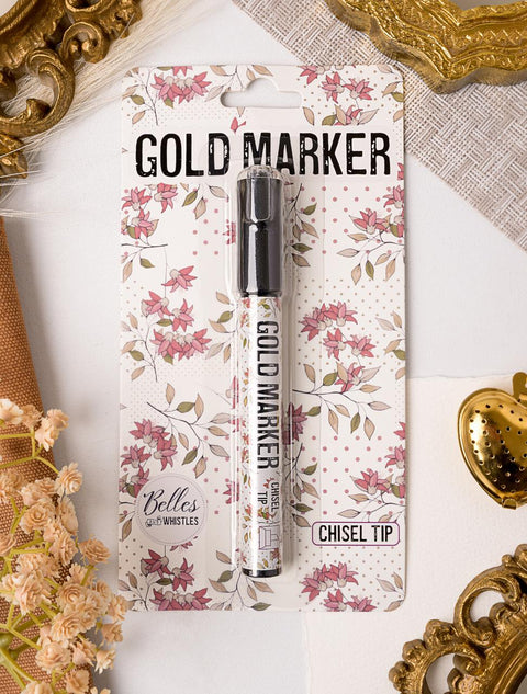 Packaging of a gold marker with a chisel tip. The card backing features a floral design in pink and white tones. The brand name "Belles and Whistles" is visible. Gold decorative elements and white fabric are in the background.