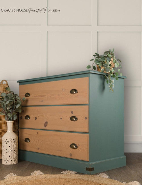 A blue-green and natural wood chest of drawers with brass handles is adorned with cascading greenery. A woven basket and a ceramic vase with leafy branches sit beside it. The setting is a light-colored room with a textured rug on the floor.