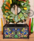 A black wooden box with intricate, colorful patterns of two birds is displayed on a wooden shelf. The box is surrounded by tall, multicolored glass bottles and a green leafy wreath is hung on a wooden frame in the background.
