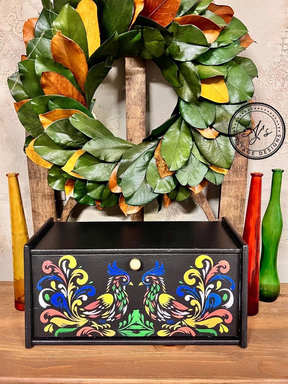A black wooden box with intricate, colorful patterns of two birds is displayed on a wooden shelf. The box is surrounded by tall, multicolored glass bottles and a green leafy wreath is hung on a wooden frame in the background.