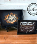 Two painted black serving trays against a white wood panel wall on a light wood floor. The larger tray to the left has the spiderweb "Happy Halloween" design applied in orange and white. The smaller tray to the right has the bat and chain Happy Halloween design applied in white and gold. One white and two orange pumpkins sit next to the trays.