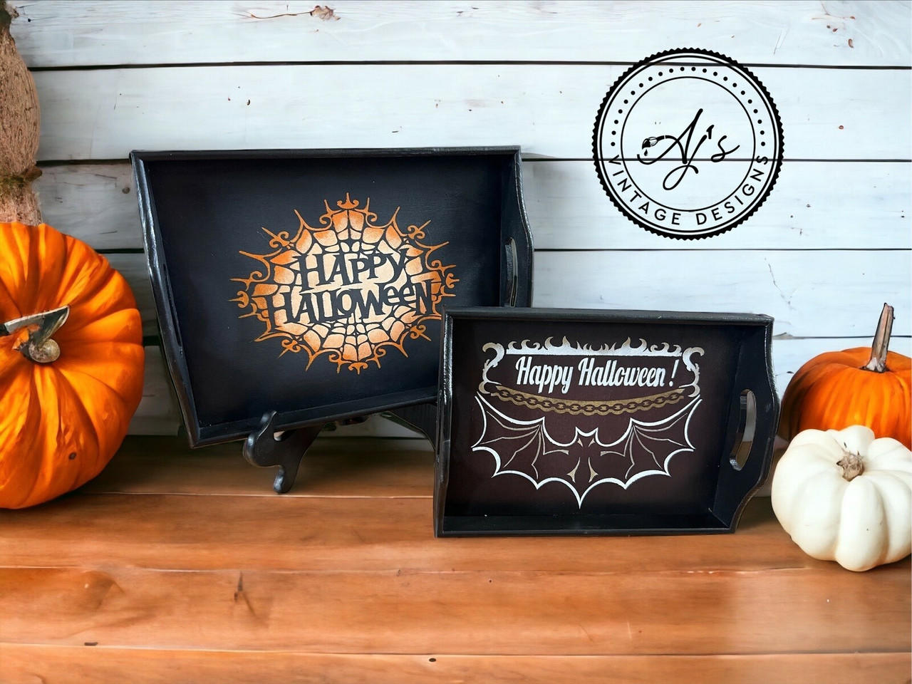 Two painted black serving trays against a white wood panel wall on a light wood floor. The larger tray to the left has the spiderweb "Happy Halloween" design applied in orange and white. The smaller tray to the right has the bat and chain Happy Halloween design applied in white and gold. One white and two orange pumpkins sit next to the trays.