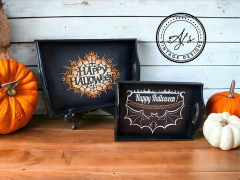 Two painted black serving trays against a white wood panel wall on a light wood floor. The larger tray to the left has the spiderweb "Happy Halloween" design applied in orange and white. The smaller tray to the right has the bat and chain Happy Halloween design applied in white and gold. One white and two orange pumpkins sit next to the trays.