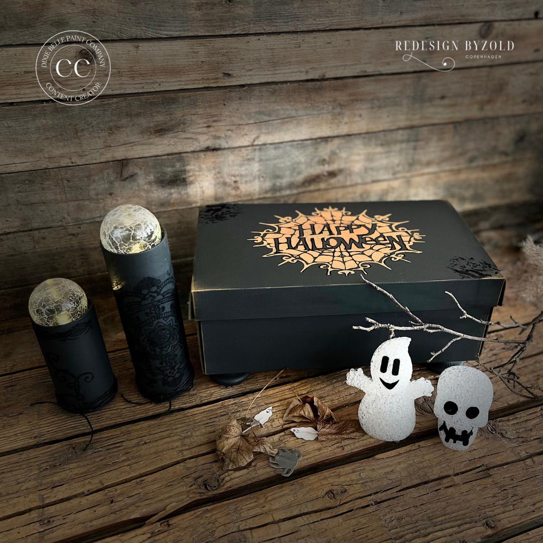 Black painted box with the spiderweb Happy Halloween Stencil design applied in goldish orange. Small white ghost figurine, small white skull figurine, two small black candle holders with orbs inside.