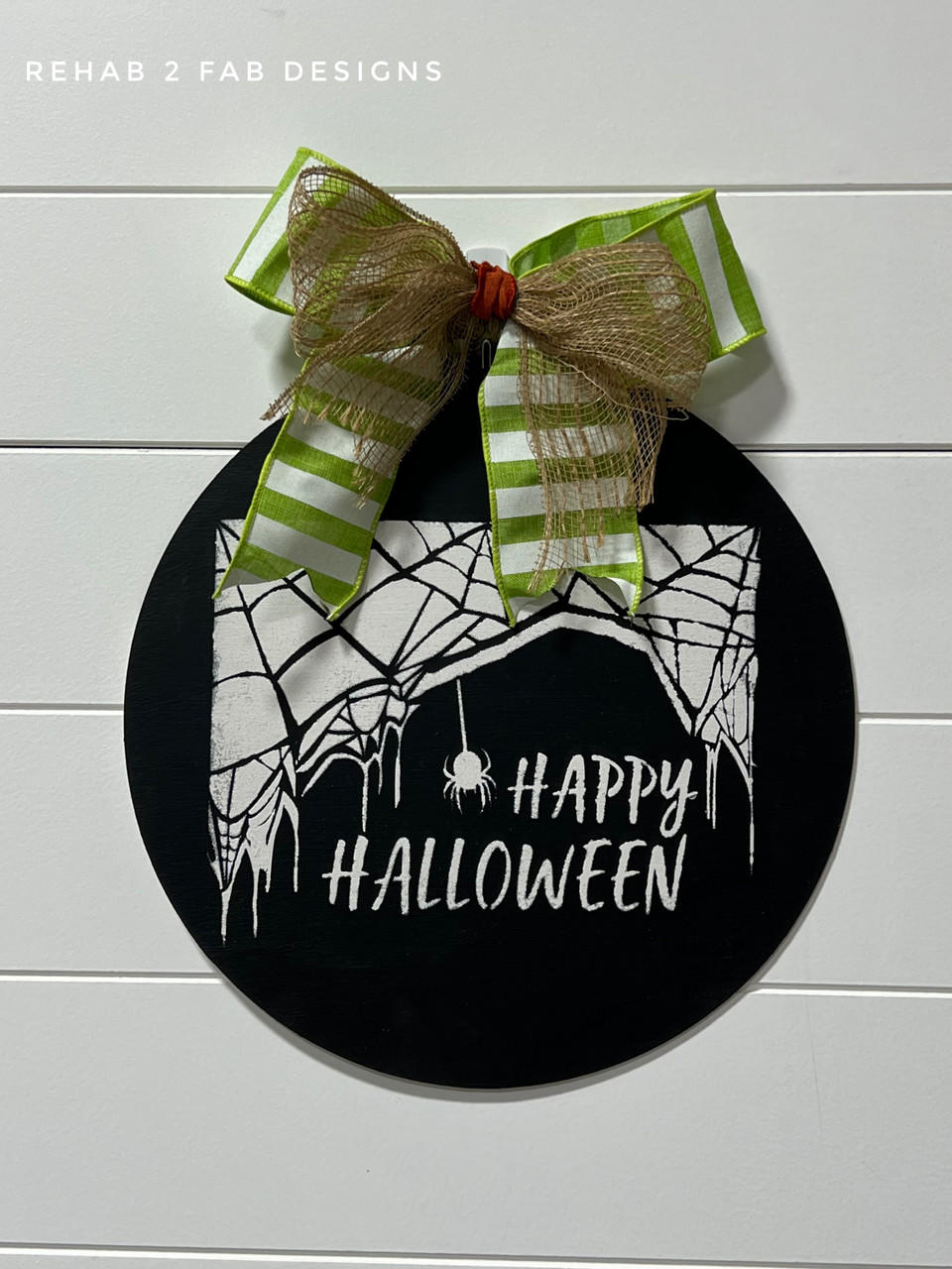 Black painted circular sign with the spiderweb and spider Happy Halloween Stencil design applied in white. Green and white striped bow on the top of the sign.