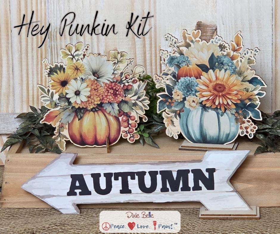 Two autumn-themed pumpkin floral arrangements and a white wooden arrow sign labeled "AUTUMN" are displayed on a wooden surface. Text at the top reads "Hey Punkin Kit." The background has a rustic, light wood texture.