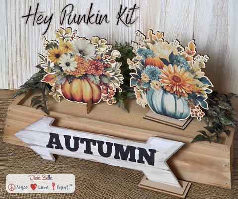 Two autumn-themed floral pumpkin decorations sit on a wooden box with foliage, next to a white wooden arrow sign reading "AUTUMN." Text above reads "Hey Punkin Kit."
