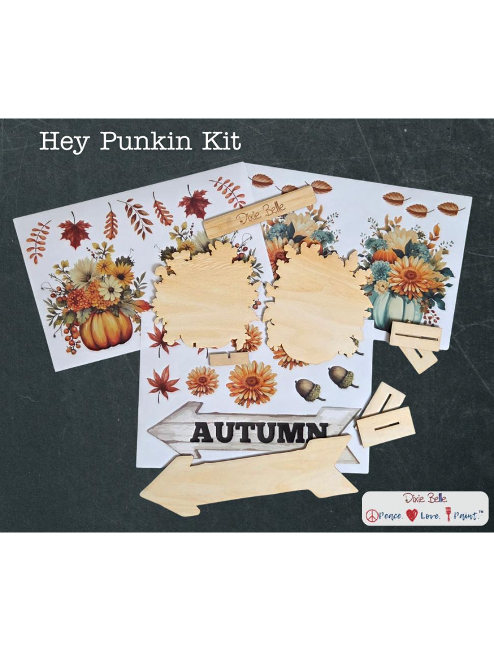 A craft kit labeled "Hey Punkin Kit" includes autumn-themed floral and pumpkin transfers, unfinished wood cutouts in various shapes, and a wooden sign reading "AUTUMN," all displayed on a dark background.