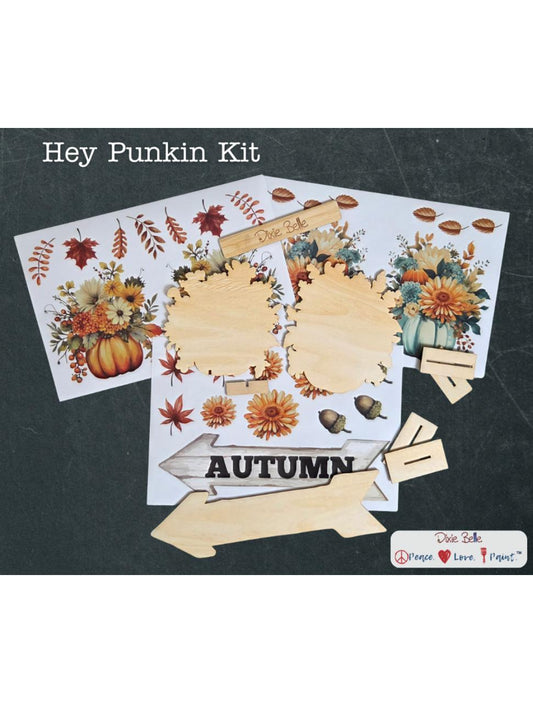 A craft kit labeled "Hey Punkin Kit" includes autumn-themed floral and pumpkin transfers, unfinished wood cutouts in various shapes, and a wooden sign reading "AUTUMN," all displayed on a dark background.