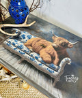 A decorative table featuring an image of a brown highland cow lounging on a blue and white floral couch, with antlers, a blue vase, and branches displayed on top. The corner reads "Tracey's Fancy."