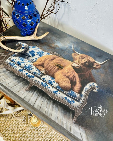 A decorative table featuring an image of a brown highland cow lounging on a blue and white floral couch, with antlers, a blue vase, and branches displayed on top. The corner reads "Tracey's Fancy."