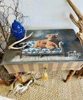 A painted side table features an image of a Highland cow lounging on a blue and white floral sofa. Decor includes a blue vase, antlers, and twigs, all set on a woven rug. "Tracey's Fancy" is written on the tabletop.