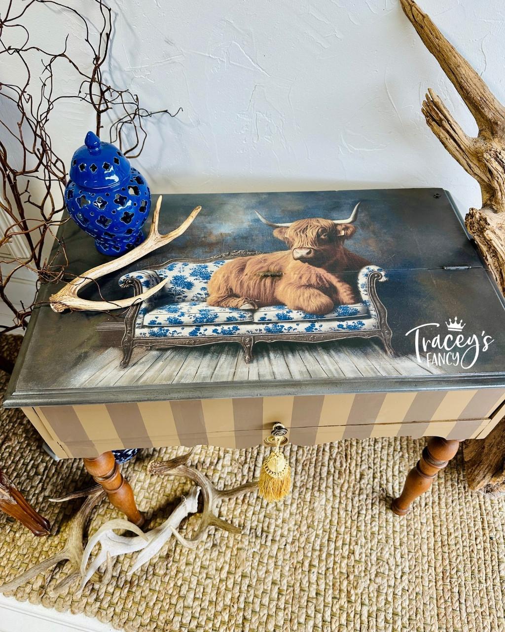 A painted side table features an image of a Highland cow lounging on a blue and white floral sofa. Decor includes a blue vase, antlers, and twigs, all set on a woven rug. "Tracey's Fancy" is written on the tabletop.