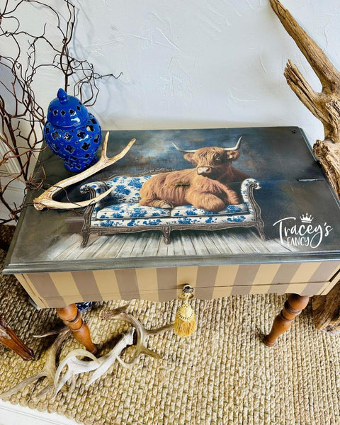A painted side table features an image of a Highland cow lounging on a blue and white floral sofa. Decor includes a blue vase, antlers, and twigs, all set on a woven rug. "Tracey's Fancy" is written on the tabletop.
