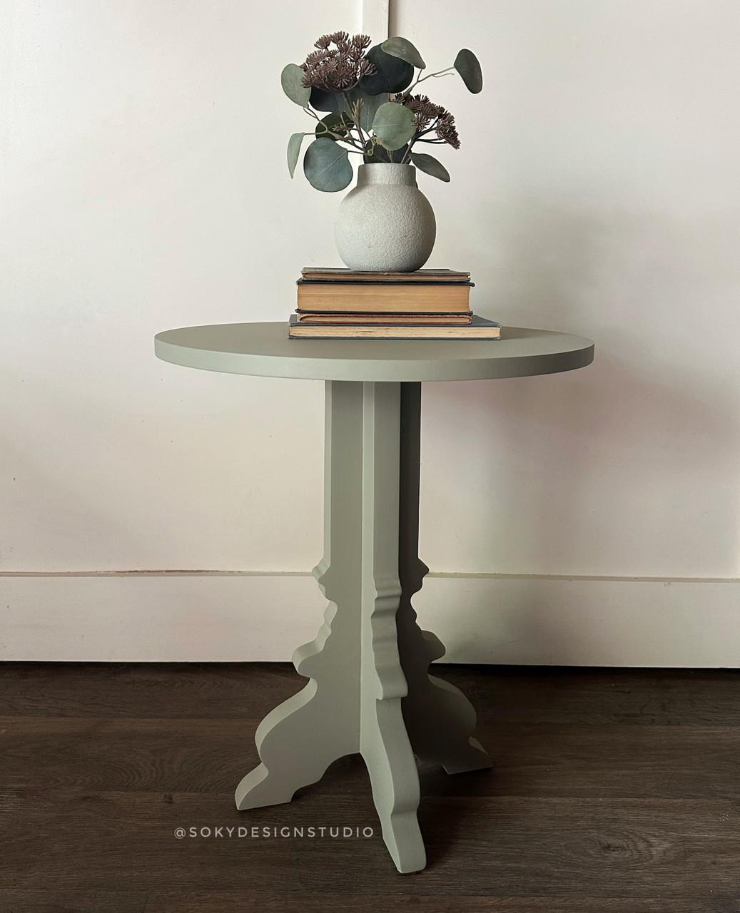 A small round table with a pale green finish holds a stack of books and a white vase filled with eucalyptus branches and dried flowers, set against a plain white wall and dark wood floor.