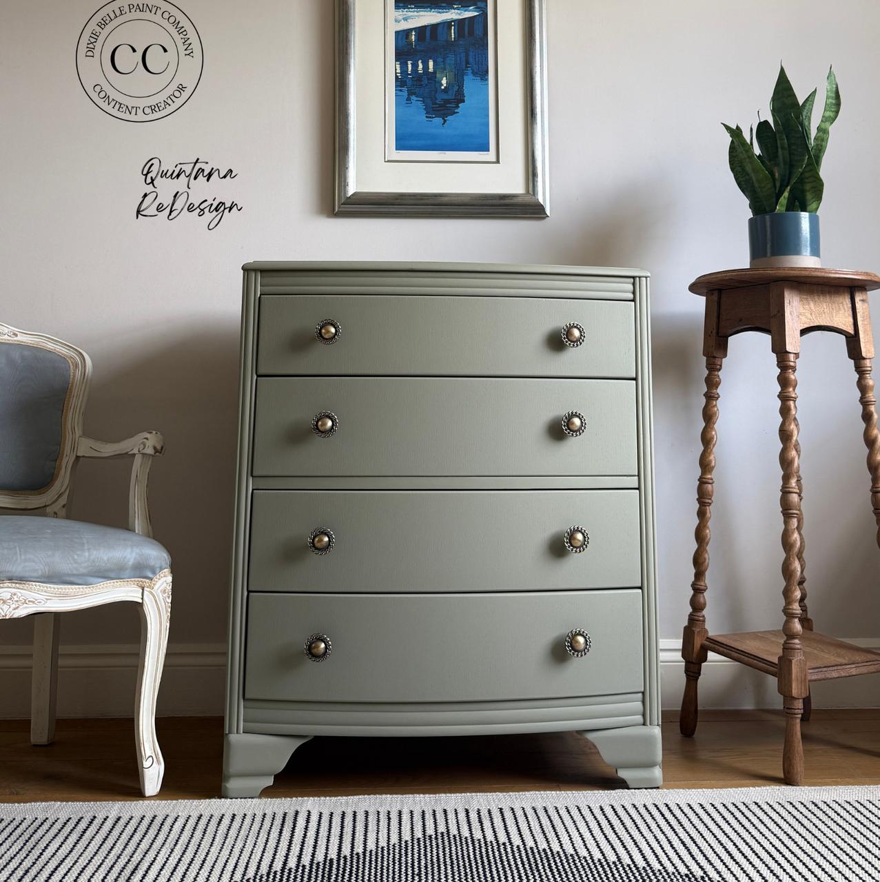 A sage green dresser with four drawers sits between a white chair and a tall wooden plant stand. A framed blue cityscape artwork hangs above, and a potted plant is on the stand. The room has neutral walls and wooden floors.