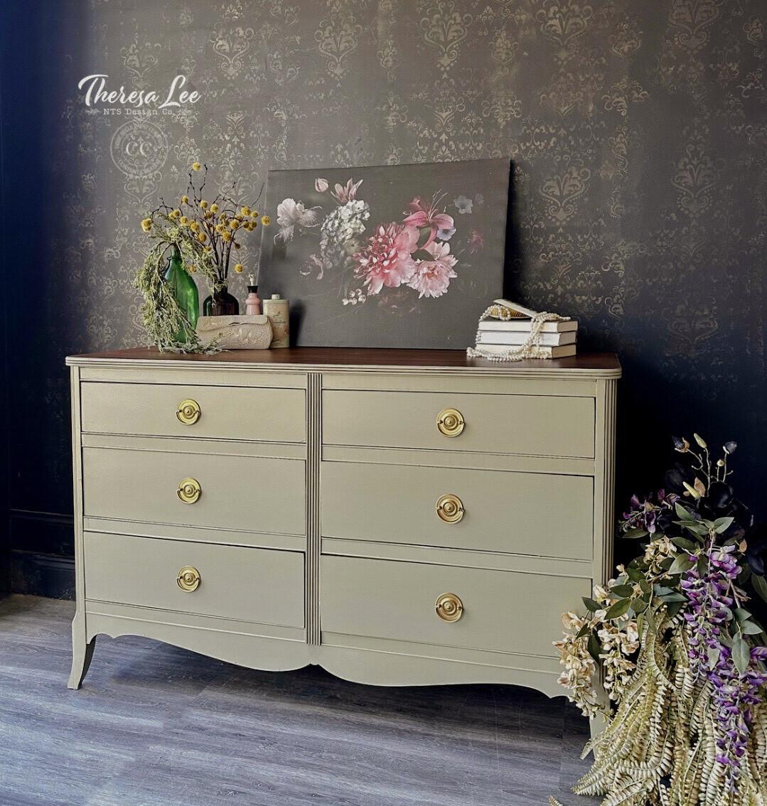A light olive six-drawer dresser with gold knobs stands against a dark, patterned wall. On top are flowers in vases, a floral painting, and stacked books. Cascading flowers spill from the dresserâ€™s side onto the floor.