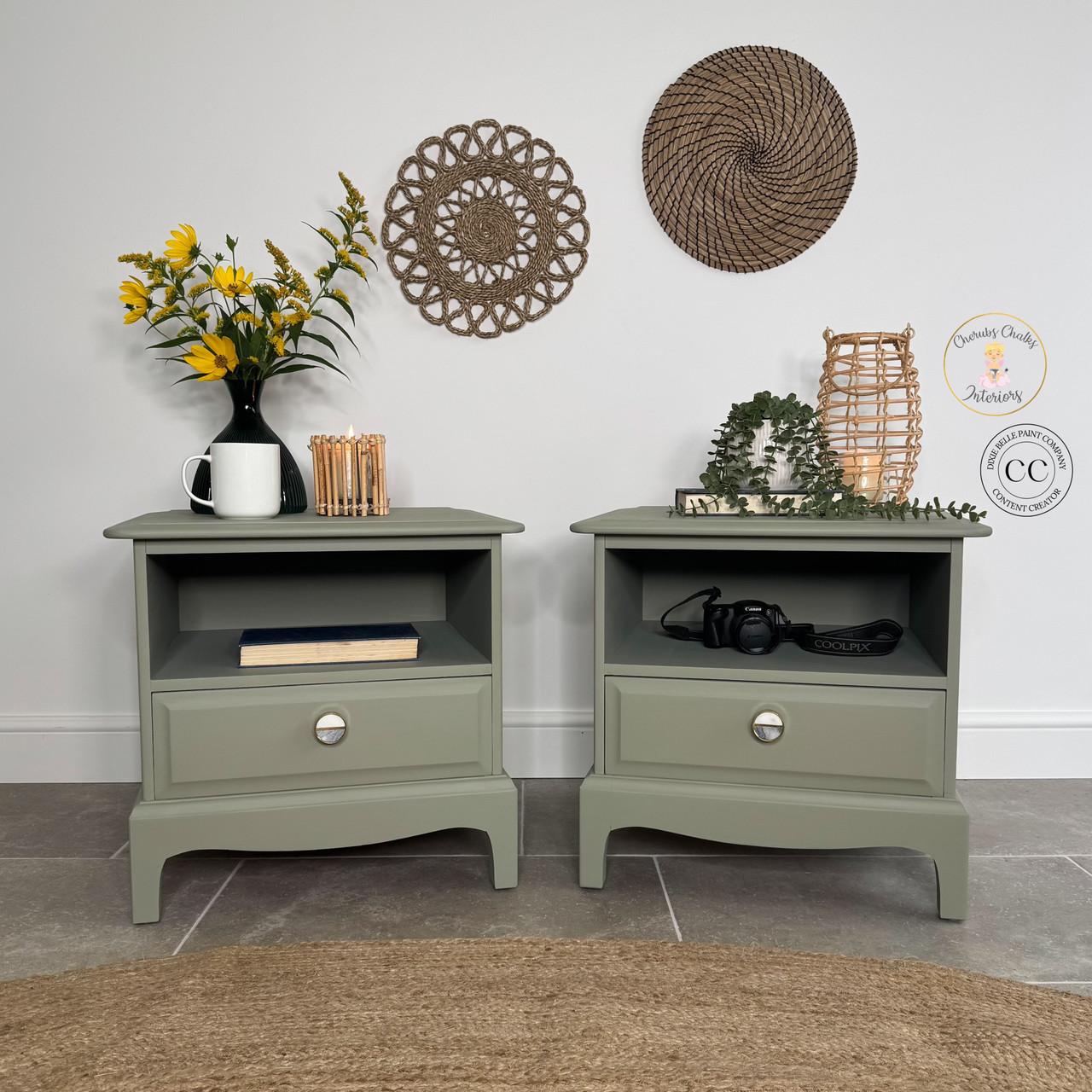 Two green nightstands with silver knobs sit side by side against a white wall, decorated with yellow flowers in a white vase, a camera, and books. Woven wall hangings are above them. A round jute rug is on the floor.