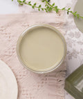 A jar filled with smooth, pale green paint sits on a textured pink cloth, surrounded by greenery and ceramic plates.