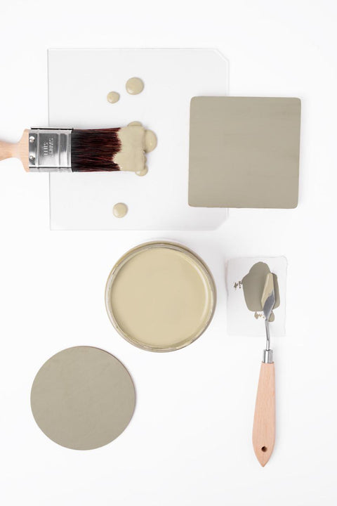 Flat lay of a paintbrush with green paint, a glass sheet with green paint drops, an open can of green paint, two green color samples, and a palette knife with green paint on a white background.