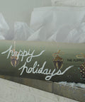 A wooden box with rope handles is filled with white tissue paper. The box displays festive decorations with pinecones and a handwritten script that reads "Happy Holidays." Snow-like decorations are scattered around the box on a white surface.