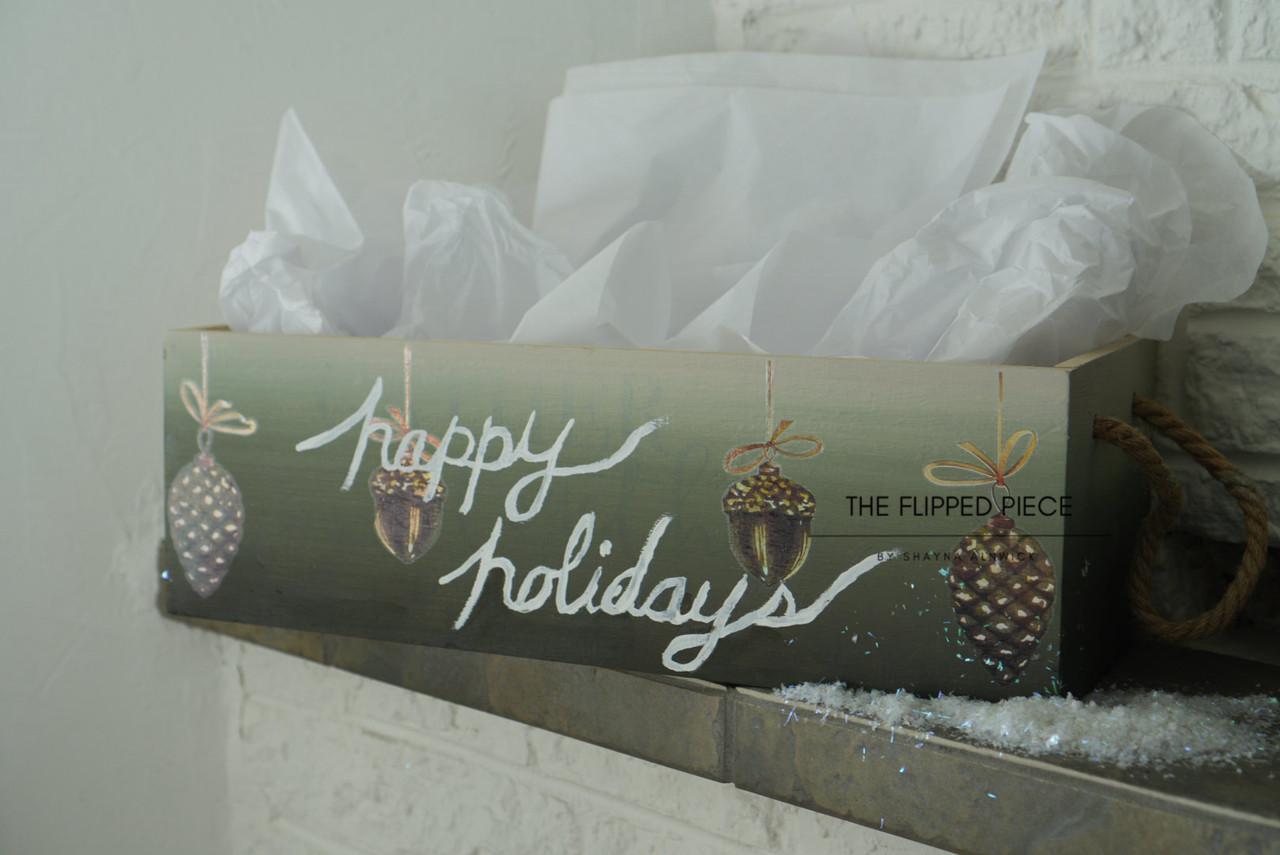 A wooden box with rope handles is filled with white tissue paper. The box displays festive decorations with pinecones and a handwritten script that reads "Happy Holidays." Snow-like decorations are scattered around the box on a white surface.