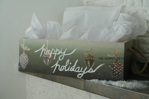 A wooden box with rope handles is filled with white tissue paper. The box displays festive decorations with pinecones and a handwritten script that reads "Happy Holidays." Snow-like decorations are scattered around the box on a white surface.