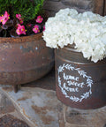 An outdoor setting featuring rustic planters with vibrant pink and white blooms. A planter with "home sweet home" painted in white on it sits beside a metal pot on a stone path.