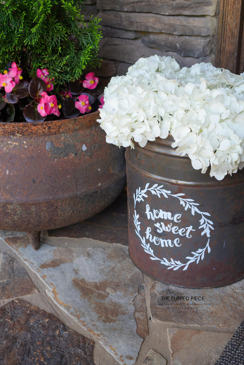 An outdoor setting featuring rustic planters with vibrant pink and white blooms. A planter with "home sweet home" painted in white on it sits beside a metal pot on a stone path.