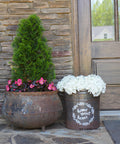 A charming entrance with a rustic metal pot and a decorative bucket, filled with pink and white flowers respectively, beside a topiary on a stone-clad porch. A reflection of a house is visible in the door’s window.