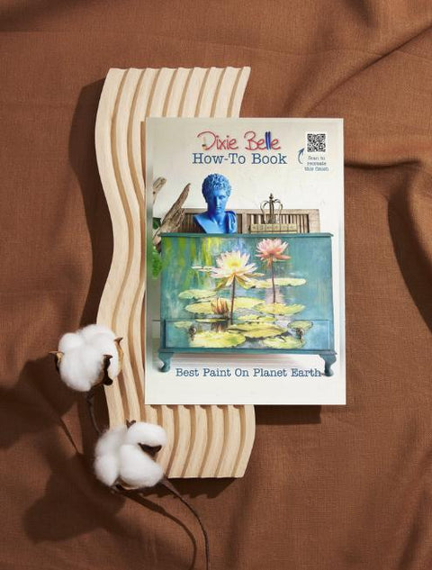 A book titled "Dixie Belle How-To Book" with a colorful cover featuring a decorative piece on a wooden wavy tray. Two cotton balls are placed beside the book on a brown fabric background.