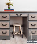 A wooden desk with six drawers is painted in gray tones labeled "Up In Smoke," accompanied by a wooden stool. On the desk, there's a vase with flowers and three stacked books.