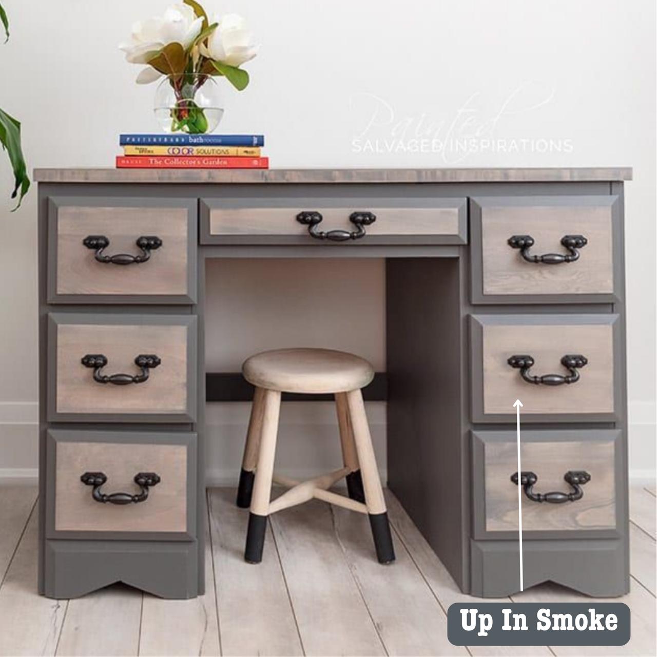 A wooden desk with six drawers is painted in gray tones labeled "Up In Smoke," accompanied by a wooden stool. On the desk, there's a vase with flowers and three stacked books.