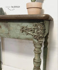 Close-up of a vintage wooden table with ornate floral carvings on its legs and edges. The table is painted in distressed green. A small terracotta pot with a plant sits on the table corner. The words "by ZOLD DESIGN" are visible on the wall.