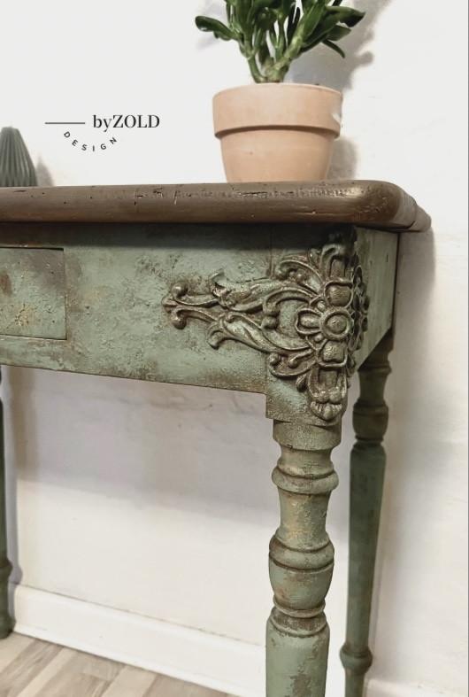 Close-up of a vintage wooden table with ornate floral carvings on its legs and edges. The table is painted in distressed green. A small terracotta pot with a plant sits on the table corner. The words "by ZOLD DESIGN" are visible on the wall.