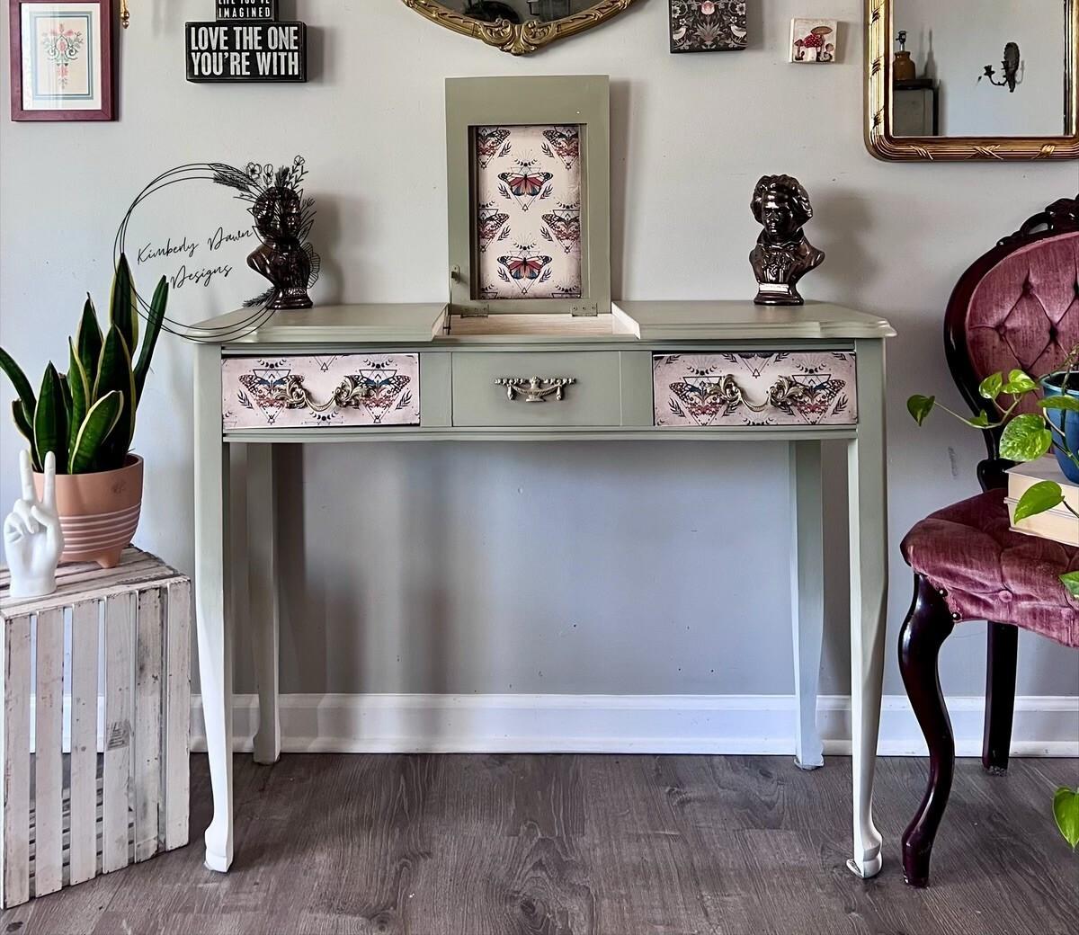 A vintage light green vanity with floral and butterfly-patterned drawers, flanked by two bust sculptures. A circular frame and various decorations are on the wall. A pink chair and potted plants complete the stylish setup.