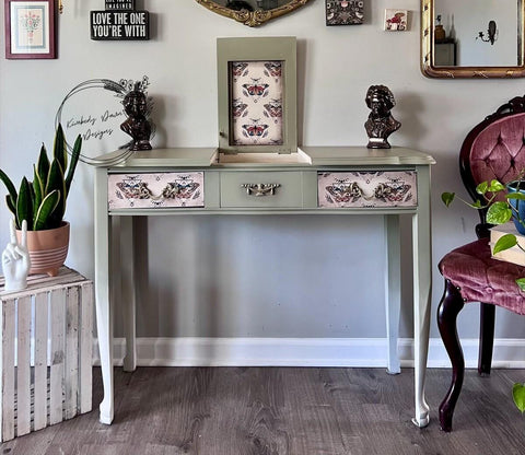 A vintage light green vanity with floral and butterfly-patterned drawers, flanked by two bust sculptures. A circular frame and various decorations are on the wall. A pink chair and potted plants complete the stylish setup.