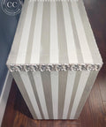 A striped rectangular chest with alternating light gray and white vertical stripes, decorative silver floral trim along the top edge, placed on a dark wooden floor next to a blue wall.